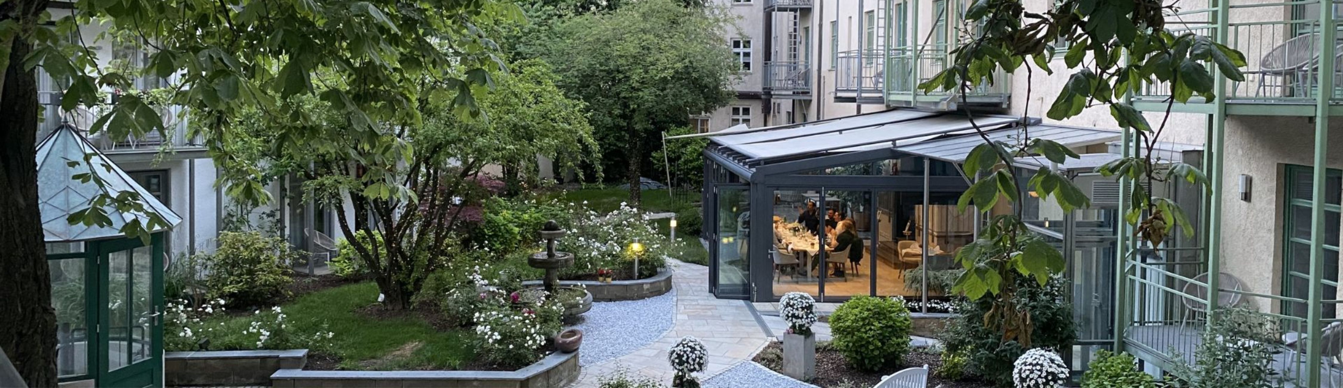 Grüner Innenhof eines Wohn- oder Hotelgebäudes mit Bäumen, Blumenbeeten und einem Glasanbau, in dem Menschen an einem gedeckten Tisch sitzen.